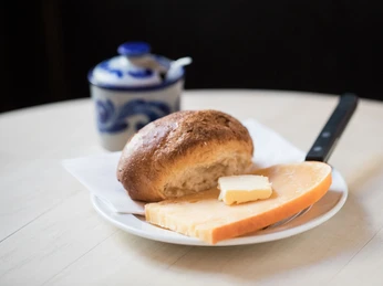 Max Stark Ein knuspriges Brötchen mit einem Stück Butter darauf liegt neben einer großzügigen Scheibe Goudakäse. A crispy roll with a pat of butter on top lies next to a generous slice of Gouda cheese.