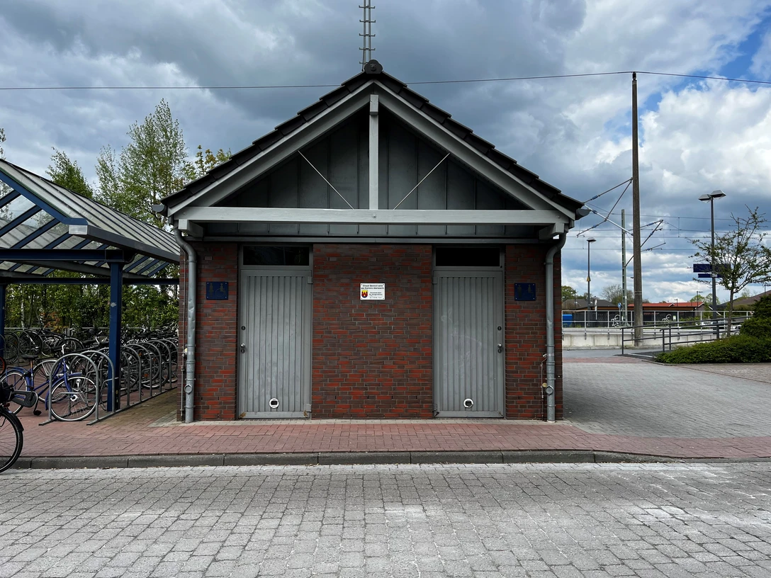 Öffentliche Toilette am Bahnhof.jpg Modernes, symmetrisches Ziegelgebäude am Bahnhof mit zwei getrennten Eingängen und asphaltiertem Vorplatz.
