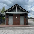 Öffentliche Toilette am Bahnhof.jpg Modernes, symmetrisches Ziegelgebäude am Bahnhof mit zwei getrennten Eingängen und asphaltiertem Vorplatz.