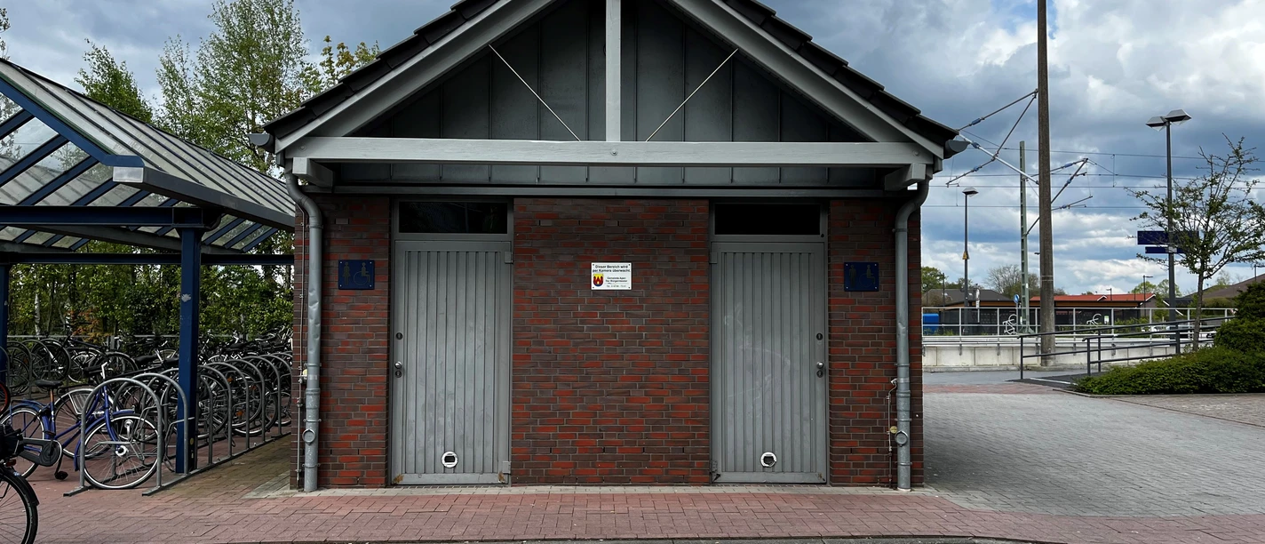 Öffentliche Toilette am Bahnhof.jpg Modernes, symmetrisches Ziegelgebäude am Bahnhof mit zwei getrennten Eingängen und asphaltiertem Vorplatz.