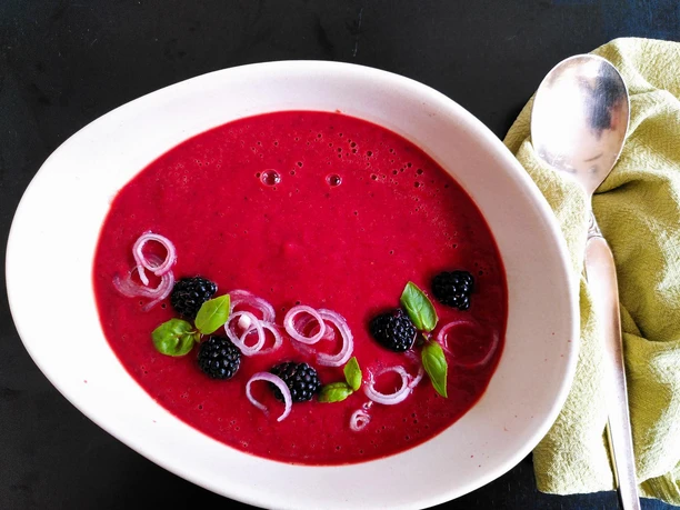 Brombeer-Gazpacho Stilvolle Schale mit leuchtend roter Suppe, garniert mit Brombeeren, Zwiebelringen und Basilikumblättern.