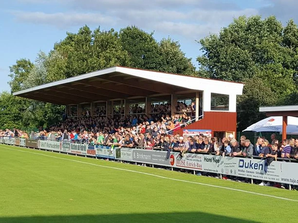 Sportplatz_TuRa_ 07_ Rhauderfehn Zuschauer sitzen auf überdachter Tribüne neben einem grünen Fußballfeld an einem sonnigen Tag