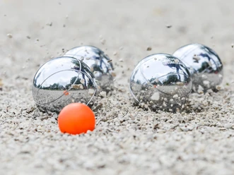 Boule-Foto-Internet.png Vier silberne Boulekugeln und eine kleine orangefarbene Zielkugel auf kiesigem Boden