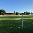 Sportanlage_Westrhauderfehn_Verein TuRa_07 Fußballfeld mit Eckfahne im Vordergrund, Zuschauertribüne und Bäume im Hintergrund bei sonnigem Wetter.