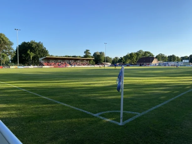 Sportanlage_Westrhauderfehn_Verein TuRa_07 Fußballfeld mit Eckfahne im Vordergrund, Zuschauertribüne und Bäume im Hintergrund bei sonnigem Wetter.