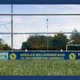 Sportanlage_SC Rhauderfehn_ Langholt Fußballtor mit Netz auf einem Spielfeld, Werbebanner mit Text 'Herzlich Willkommen beim SC Rhuderfehn Langholt' und Firmenlogos im Hintergrund.