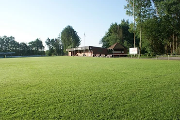 Sportanlage_SV_Burlage_e.V. Grünes Fußballfeld mit einem Klinkerbau und einer angrenzenden Holzhütte und Bäumen im Hintergrund an einem sonnigen Tag.