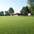 Sportanlage_SV_Burlage_e.V. Grünes Fußballfeld mit einem Klinkerbau und einer angrenzenden Holzhütte und Bäumen im Hintergrund an einem sonnigen Tag.