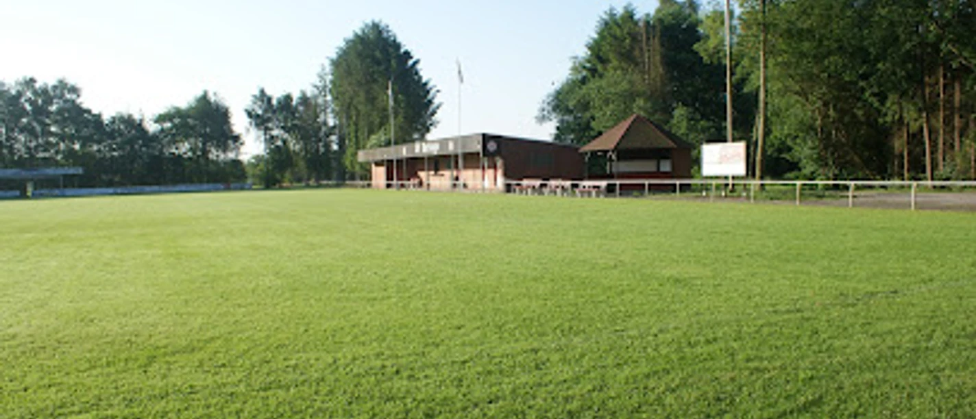 Sportanlage_SV_Burlage_e.V. Grünes Fußballfeld mit einem Klinkerbau und einer angrenzenden Holzhütte und Bäumen im Hintergrund an einem sonnigen Tag.