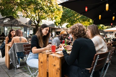 Freddy Schilling Eigelstein Junge Mensch essen Burger an einem Holztisch in der Außengastronomie.