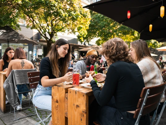 Freddy Schilling Eigelstein Junge Mensch essen Burger an einem Holztisch in der Außengastronomie.Young people eating burgers at a wooden table in an outdoor restaurant.