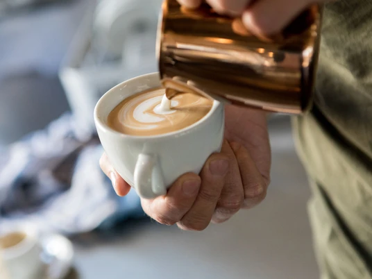 Cafe Heilandt Nahaufnahme, in eine Tasse Kaffee wird aufgeschäumte Milch gegossen.Close-up of milk being poured into a cup of coffee.