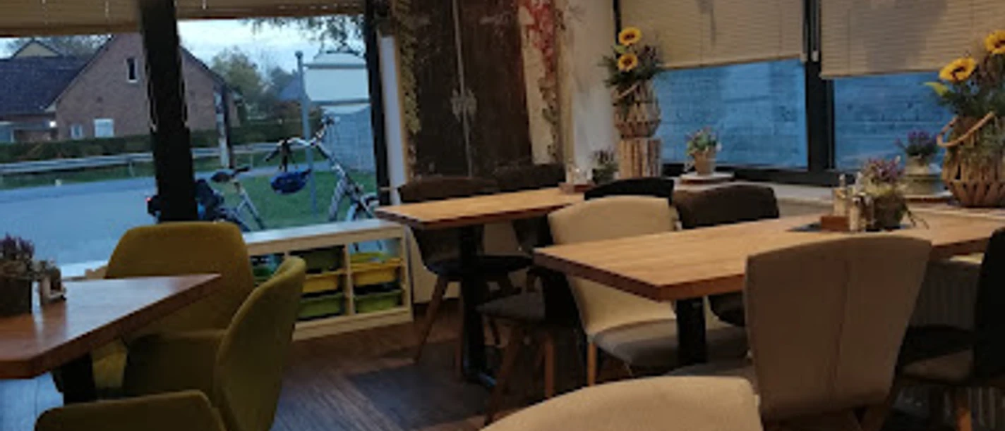 Bäckerei Ripken_Edeka_Augustfehn_2022.jpg Das Innere der Bäckerei Ripken in Augustfehn strahlt eine gemütliche und einladende Atmosphäre aus. Holzvertäfelte Möbel und dezente Dekorationen, wie Trockenblumen und moderne Lampen, schaffen ein stilvolles, rustikales Ambiente. Große Fenster sorgen für viel Tageslicht und geben den Blick auf die Umgebung frei, während die Tischdekoration mit kleinen Kräutertöpfen den ländlichen Charme unterstreicht.