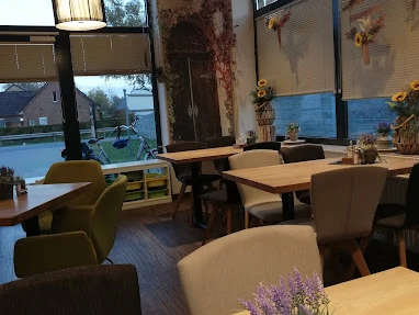 Bäckerei Ripken_Edeka_Augustfehn_2022.jpg Das Innere der Bäckerei Ripken in Augustfehn strahlt eine gemütliche und einladende Atmosphäre aus. Holzvertäfelte Möbel und dezente Dekorationen, wie Trockenblumen und moderne Lampen, schaffen ein stilvolles, rustikales Ambiente. Große Fenster sorgen für viel Tageslicht und geben den Blick auf die Umgebung frei, während die Tischdekoration mit kleinen Kräutertöpfen den ländlichen Charme unterstreicht.