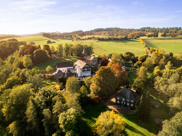 Kürten-Delling Luftaufnahme einer idyllischen, grünen Hügellandschaft mit verstreuten Gebäuden und herbstlichen Bäumen.