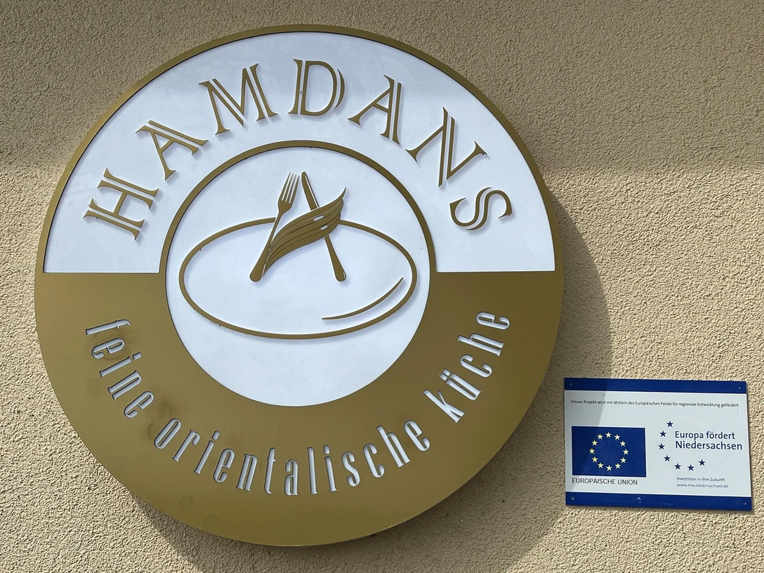Hamdans Ein rundes Schild der orientalischen Küche "Hamdans" neben einem EU-Förderprojekt-Hinweis.A round sign for the oriental cuisine "Hamdans" next to an EU funding project notice.Et rundt skilt for det orientalske køkken "Hamdans" ved siden af et opslag om et EU-finansieringsprojekt.Een rond bord voor de oosterse keuken "Hamdans" naast een aankondiging van een EU-subsidieproject.