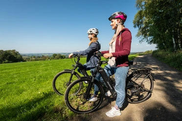 Heckberg Zwei Radfahrer mit Helmen genießen die Aussicht auf eine grüne, hügelige Landschaft bei Sonnenschein.