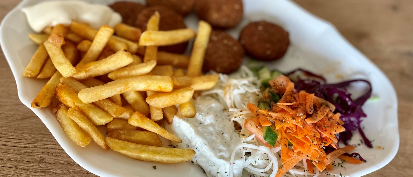 Ein Teller mit Pommes, Falafel, Krautsalat und Saucen auf einem holzfarbenen Untergrund.