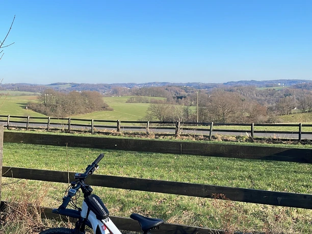 Radfahren in Nümbrecht Ein weißes Fahrrad steht vor einem Holzzaun, mit weitem Blick über Felder und blauen Himmel.