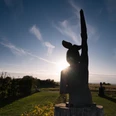 Eine Holzskulptur erhebt sich vor einem strahlenden Sonnenuntergang, umgeben von einer Wiesenlandschaft.