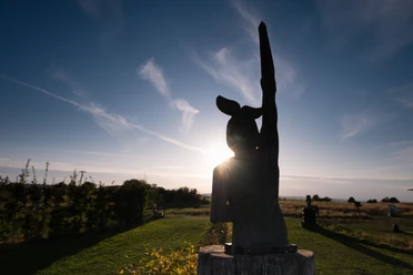 Soccerpark Detmold Eine Holzskulptur erhebt sich vor einem strahlenden Sonnenuntergang, umgeben von einer Wiesenlandschaft.