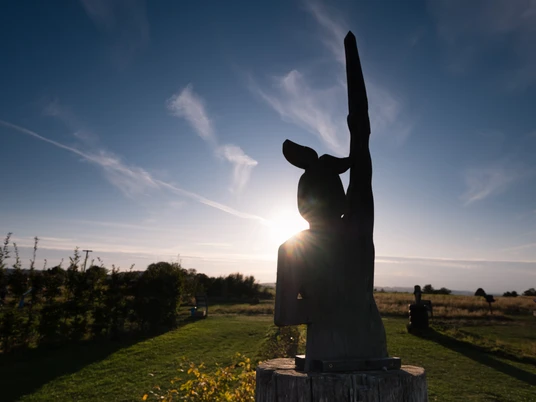 Soccerpark Detmold Eine Holzskulptur erhebt sich vor einem strahlenden Sonnenuntergang, umgeben von einer Wiesenlandschaft.
