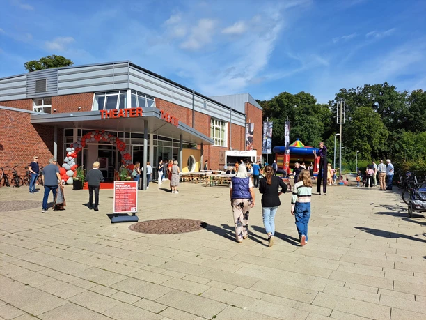Menschen besuchen den Eröffnungstag am Theater Meppen mit Ballonbogen, Stelzenläufer und Sonne.