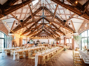 26 - Schloss_Arff_26.jpg Stilvolle Eventscheune mit großen Fenstern, hellen Holzmöbeln in langen, gedeckten Tischreihen und Lichterketten an der DeckeStylish event barn with large windows, light wooden furniture arranged in long, covered rows of tables and fairy lights hanging from the ceiling.