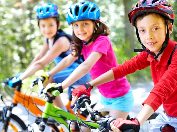 bad-driburg_kinderfahrrad_öffentlich frei.jpg Drei fröhliche Kinder fahren mit farbenfrohen Helmen auf ihren Fahrrädern durch einen sonnigen Park.