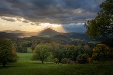 Mountain landscape at sunset with dramatic clouds, sunbeams, green meadows and colorful trees in the foreground.