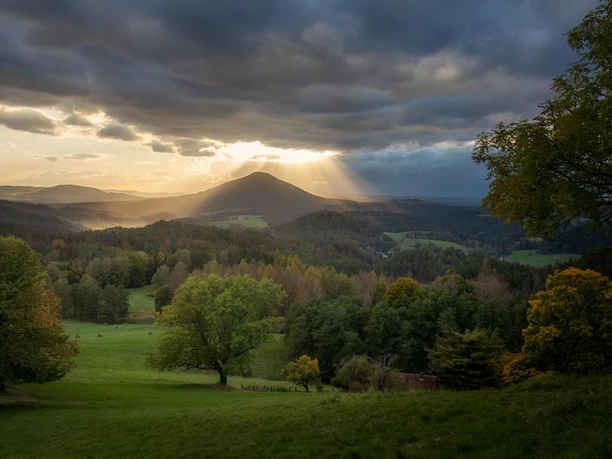 Berglandschap bij zonsondergang met dramatische wolken, zonnestralen, groene weiden en kleurrijke bomen op de voorgrond.