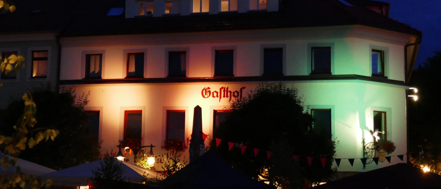 Hilligs bei Nacht Ein beleuchtetes Gasthofgebäude bei Nacht, mit Menschen unter bunten Lichtern davor.