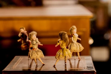 Drehorgelmuseum Vier antike Puppen in viktorianischer Kleidung stehen auf einem Holztisch in warmem Licht.