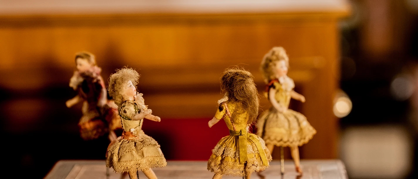 Drehorgelmuseum Vier antike Puppen in viktorianischer Kleidung stehen auf einem Holztisch in warmem Licht.
