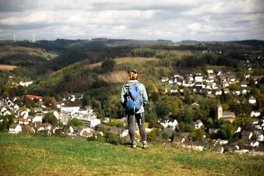 Panorama Eine Wanderin mit Rucksack blickt von einem grünen Hügel auf ein malerisches Dorf im Tal.