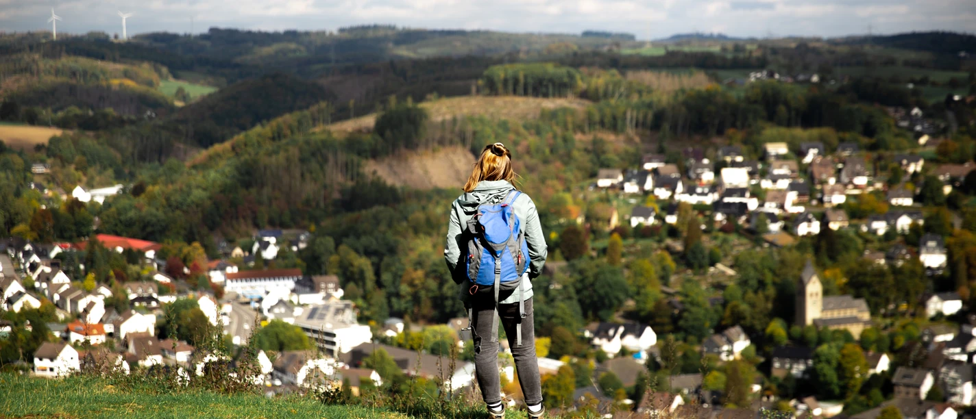 Panorama Eine Wanderin mit Rucksack blickt von einem grünen Hügel auf ein malerisches Dorf im Tal.