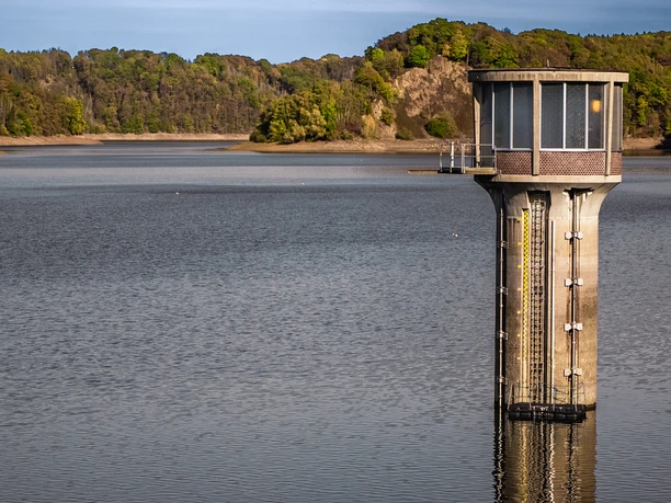 Ein grauer Wasserturm ragt aus einem ruhigen, von grünen Hügeln umgebenen Gewässer hervor.