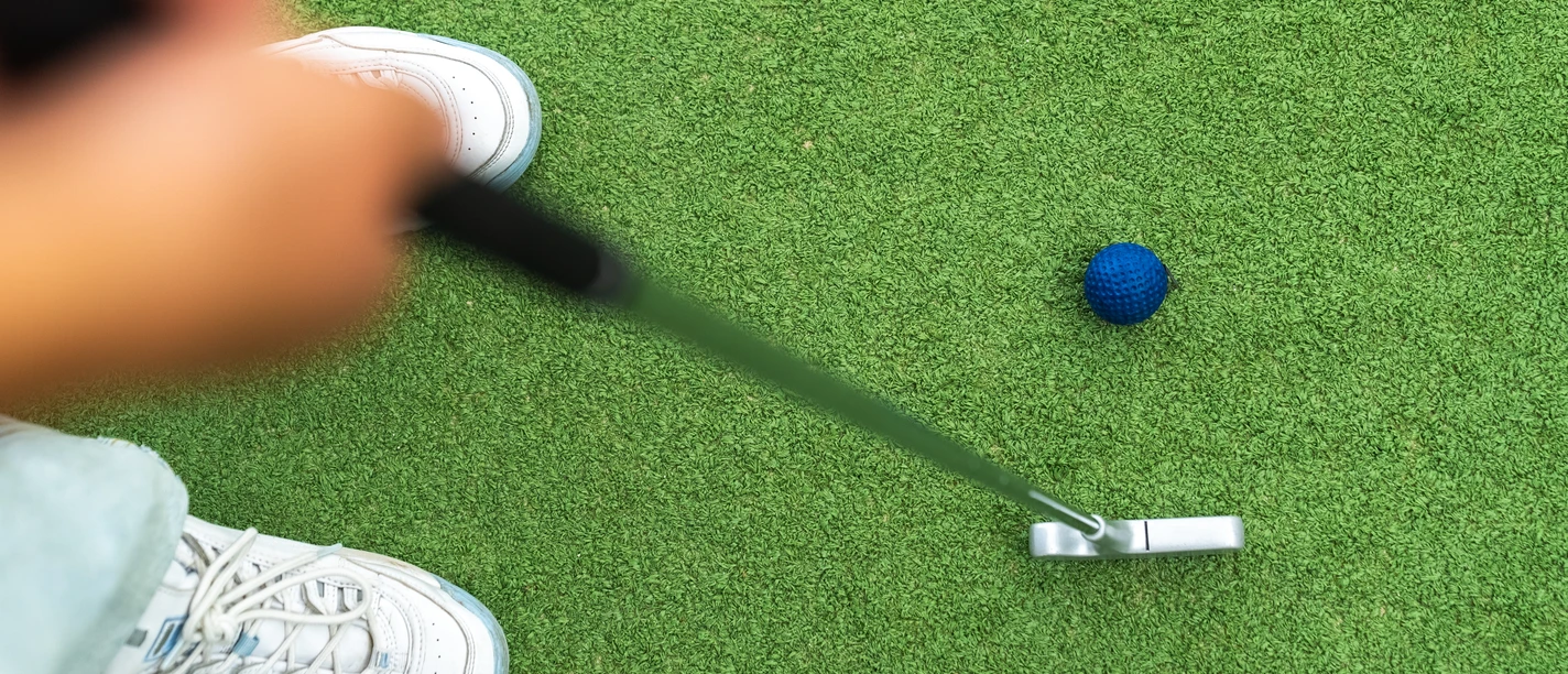 Adventure Golf Minigolfspieler trifft mit einem Putter auf einen blauen Ball auf grünem Kunstrasen.