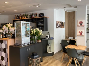 nostimo_2.jpg Gastronomieinnenraum mit Tischen und Stühlen in grau, weiß, schwarz und Holzoptik, kleine Vitrine mit Auslage im Vordergrund