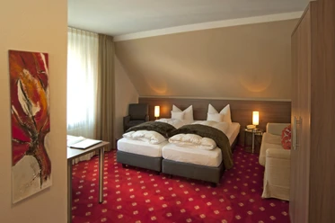 Doppelzimmer Isselhorster Landhaus Gemütliches Hotelzimmer mit zwei Einzelbetten, rotem Teppich, modernem Kunstwerk und sanfter Beleuchtung.