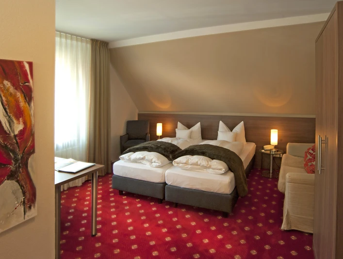 Doppelzimmer Isselhorster Landhaus Gemütliches Hotelzimmer mit zwei Einzelbetten, rotem Teppich, modernem Kunstwerk und sanfter Beleuchtung.