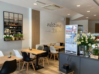 nostimo_4.jpg Gastronomieinnenraum mit Tischen und Stühlen in grau, weiß, schwarz und Holzoptik, kleine Vitrine mit Auslage im Vordergrund