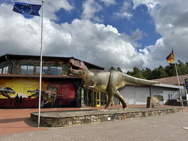 Dinopark Münchehagen Ein Dinosauriermodell vor dem Dinopark Münchehagen, umgeben von Flaggen und bewölktem Himmel.