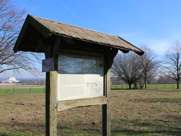 Stadt Rehburg-Loccum Kloster Asbeke Ein hölzernes Informationsschild im Freien vor einer weitläufigen grünen Wiesenlandschaft.