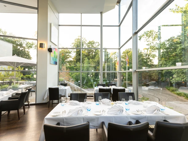 Restaurant Jordans Das Bild zeigt die moderne Innenansicht des Restaurants Jordans mit großen Glasfenstern, die viel Tageslicht hereinlassen und einen Blick ins Grüne ermöglichen. Die Einrichtung besteht aus eleganten schwarzen Stühlen und weißen Tischen, die in einem offenen und stillvollen Ambiente angeordnet sind.