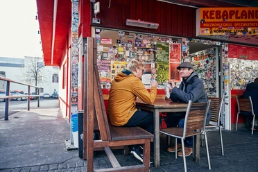 Kebapland Zwei Männer sitzen vor dem Imbiss an einem Holztisch und essen Kebap Dürüm. Die Fassade ist rot gestrichen und die Fenster mit Stickern beklebt.
