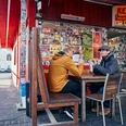 Kebapland Zwei Männer sitzen vor dem Imbiss an einem Holztisch und essen Kebap Dürüm. Die Fassade ist rot gestrichen und die Fenster mit Stickern beklebt.Two men are sitting at a wooden table in front of the snack bar eating kebap dürüm. The façade is painted red and the windows are covered with stickers.