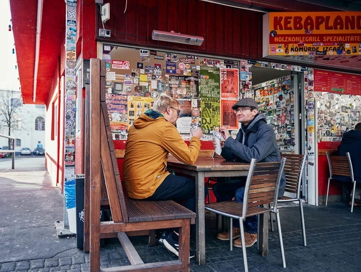 Kebapland Zwei Männer sitzen vor dem Imbiss an einem Holztisch und essen Kebap Dürüm. Die Fassade ist rot gestrichen und die Fenster mit Stickern beklebt.Two men are sitting at a wooden table in front of the snack bar eating kebap dürüm. The façade is painted red and the windows are covered with stickers.