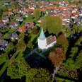 Luftaufnahme einer weißen Kirche mit grünem Turm in einem grünen, bewaldeten Dorfumfeld.