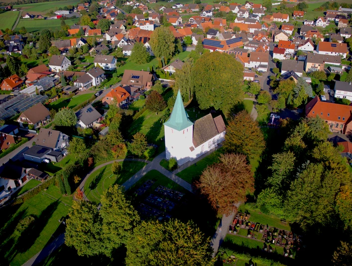 Luftaufnahme einer weißen Kirche mit grünem Turm in einem grünen, bewaldeten Dorfumfeld.